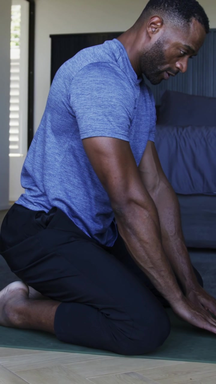 Vertical video: Sliding arms forward from heels man reaching yoga mat by bed in blue tee to stretch
