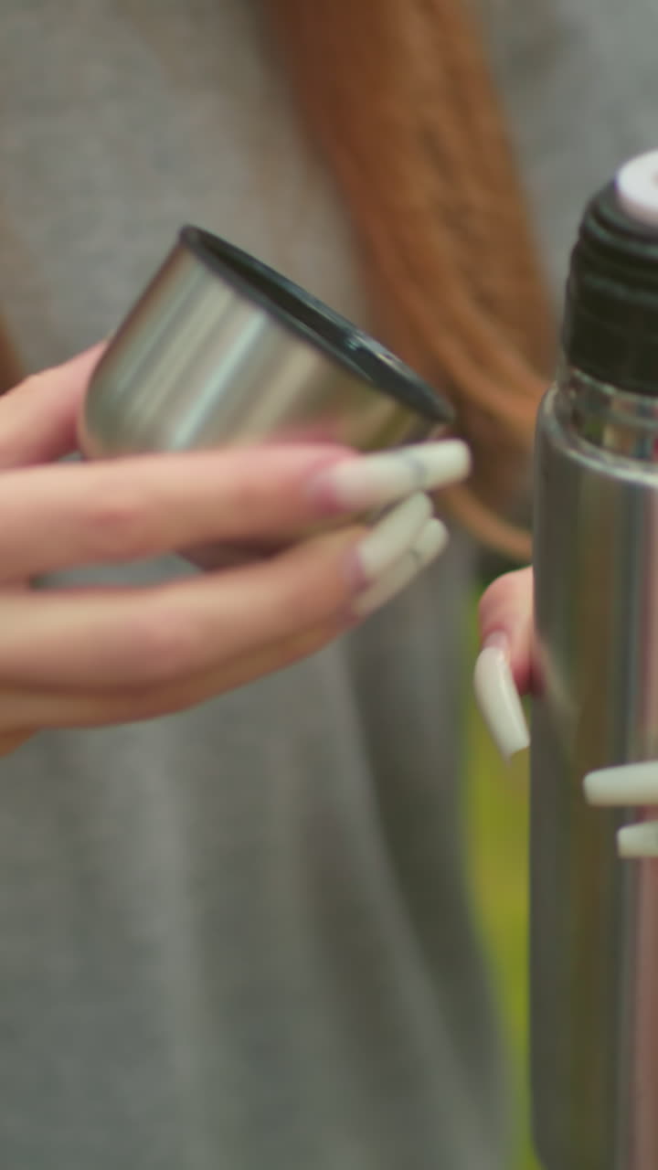 primer plano de una mujer joven con suéter gris y cabello largo cayendo en cascada, presionando la cubierta interior del termo, vertiendo agua en la tapa, destacando las uñas delicadas y largas en un entorno forestal