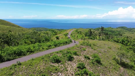 two people ride a scooter down a scenic road with ocean views and lush green hills in the distance