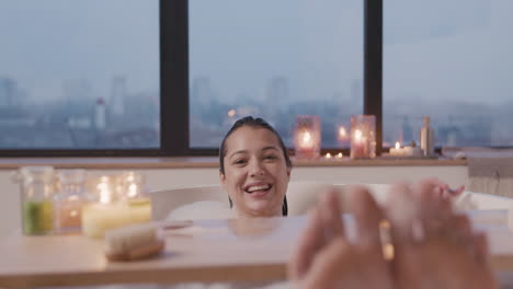 close up view of woman feet, she looking and smiling at camera while taking a bath in the bathtub decorated by a wooden table with candles