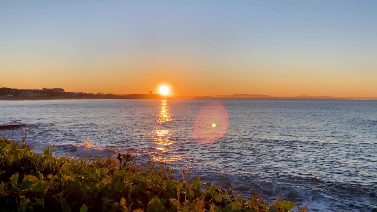 timelapse de um nascer do sol sobre o castelo na praia de carcavelos com pequenas ondas quebrando na costa e grandes reflexos na água