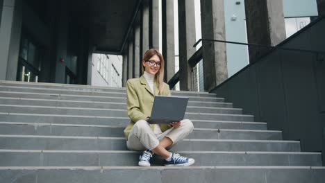 portrait of cheerful young businesswoman working online with laptop sitting on stairs outside the business center. business or education concept