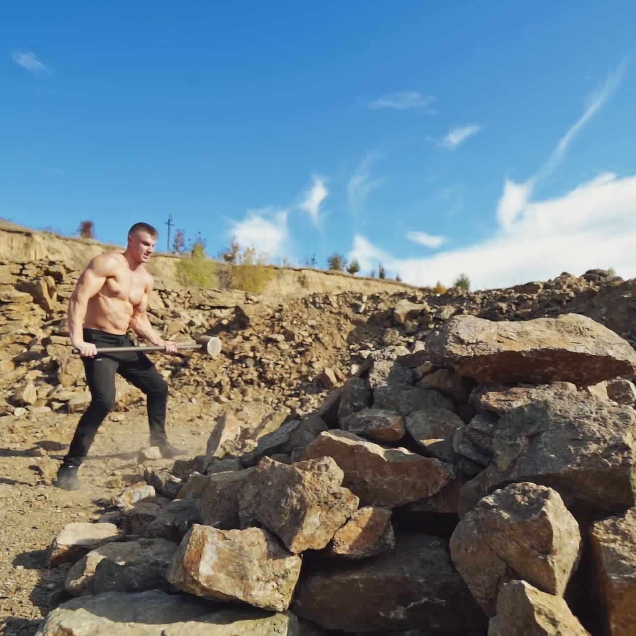 Shirtless sportsman with huge hammer outdoors. Strong athlete breaking stones with a hard steel hammer on the rocky hill background.