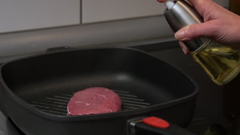 primer plano de una mujer que usa un rociador de aceite para freír un filete de ternera en una sartén caliente con aceite de oliva mientras prepara una cena deliciosa