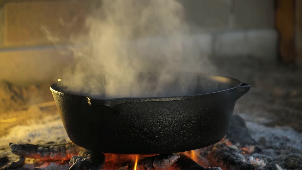 Calderone in ghisa fumante sopra le braci, pentola annerita appoggiata sul focolare, fiamme che brillano sotto un fumo denso.