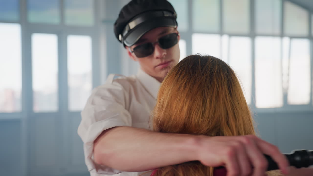 woman seated in red shirt facing mirror as man in black cap sunglasses and white shirt slides baton behind her head while dancing close in bright minimalist studio room with large windows