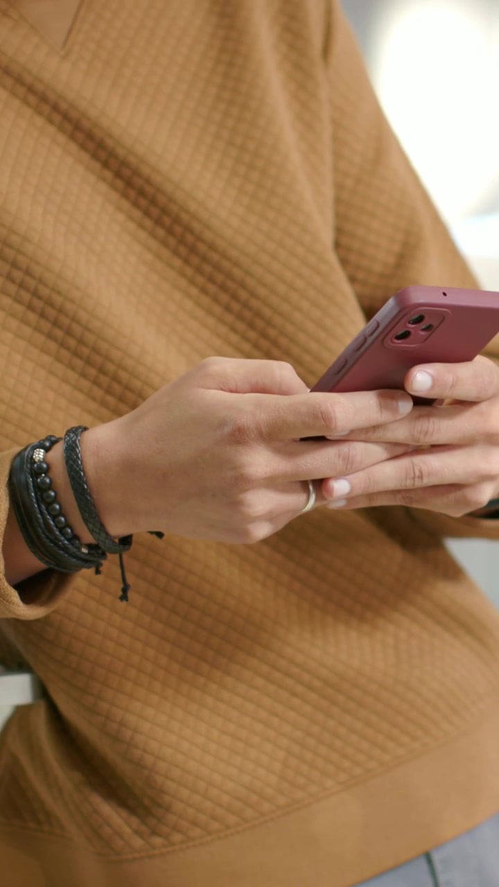 Video verticale: uomo che scorre il telefono rosa in salotto, indossa un maglione marrone, braccialetti e un anello