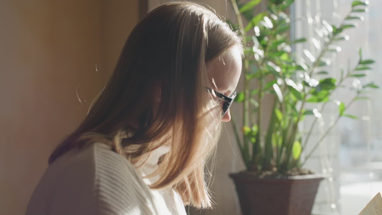 Lady in glasses reading book near window as sunlight streams in, soft focus background featuring wall art, green potted plant, coffee mug, and decorative string lights