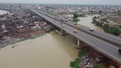 Los-Coches-Se-Mueven-Rápidamente-Sobre-El-Puente-Del-Río-Marrón-De-Una-Carretera-Suburbana-Con-Una-Bandera-Nigeriana-Frente-A-Una-Industria.