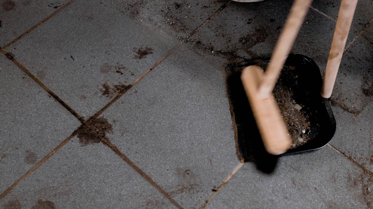 Static close up sweeping up dirt in a dust pan from a dirty tiled garage floor.