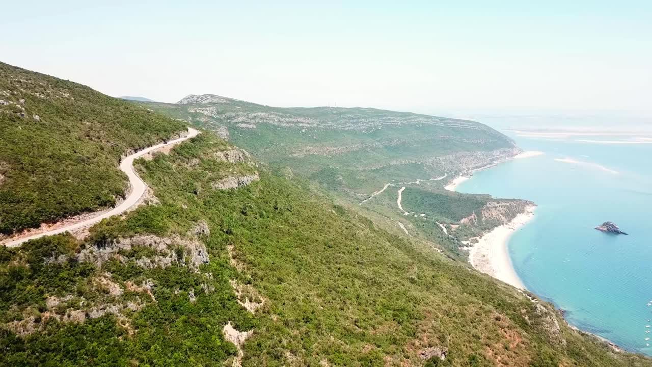 A winding road traverses lush green hills overlooking the pristine beaches and turquoise waters of Parque Natural da Arrábida, Portugal. Stunning views of rugged cliffs and serene coastal landscapes