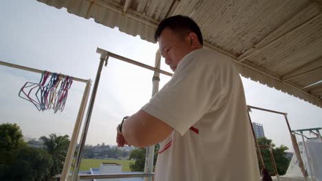 asian man checks his watch and sets a timer before hanging clothes to dry outside on his balcony - slow motion