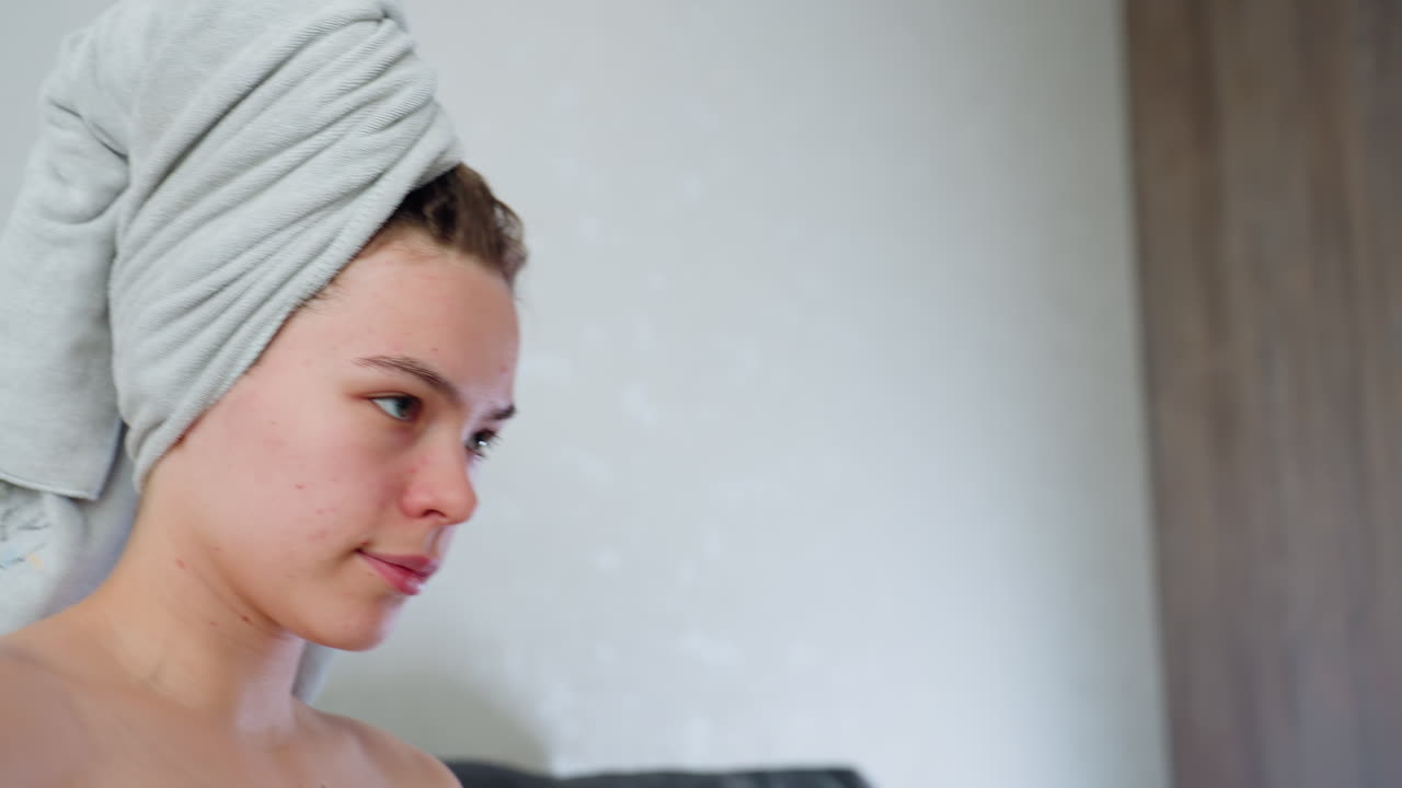 Close up side view of young woman with towel wrapped around head and green towel over chest, showing natural skin with visible acne, appearing calm and introspective in soft indoor lighting