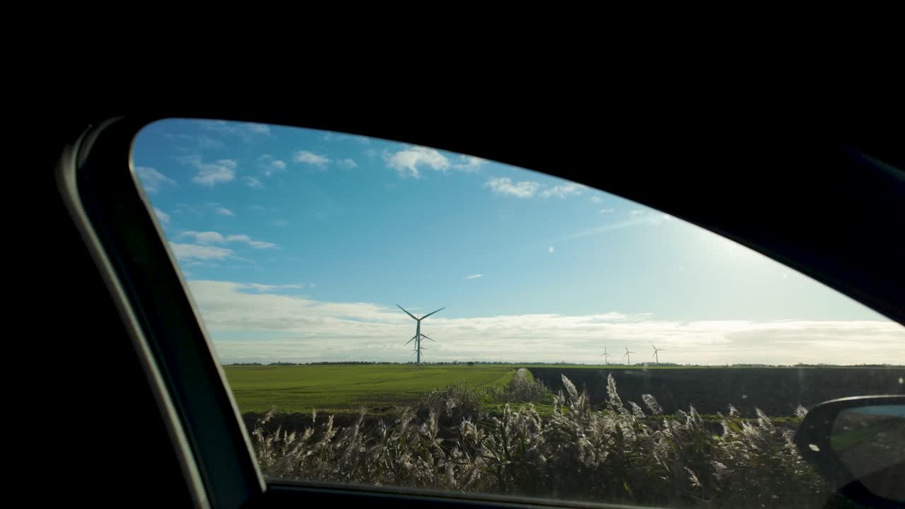 Static shot green fields with a wind turbine through a car window on a bright sunny day
