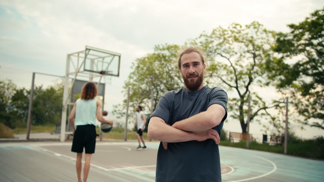 un hombre pelirrojo con una camiseta gris posa y dobla los brazos en su pecho frente a un equipo que juega baloncesto en una cancha de baloncesto de verano