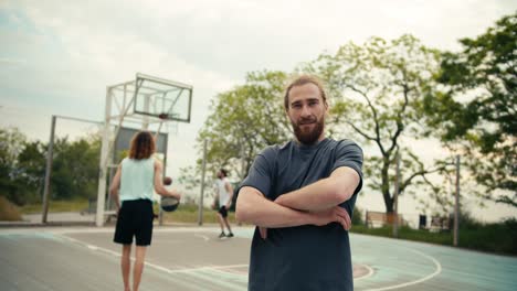 a red-haired man in a gray t-shirt poses and folds his arms on his chest in front of a team that plays basketball on a basketball court in summer