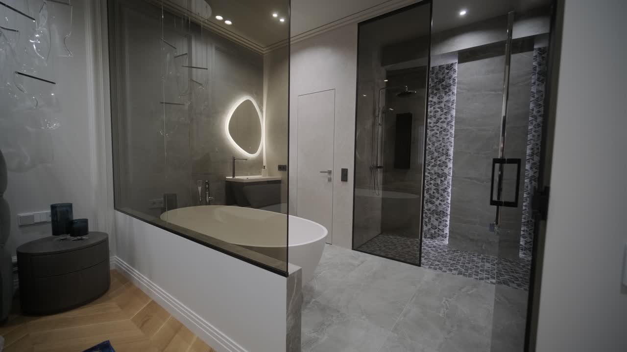 침실에서 볼 때 우아하고 현대적인 욕실, 백라이팅이 있는 타원형 거울, 독립된 욕조, 그리고 기하학 타일로 된 워크인 샤워실이 있습니다.