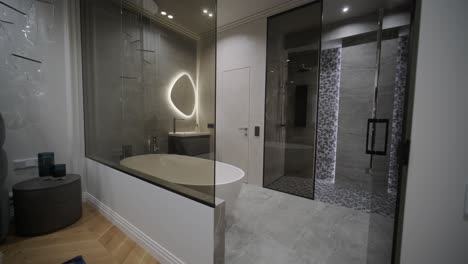 elegante en moderne badkamer gezien vanuit een slaapkamer, met een ovale spiegel met achtergrondverlichting, een vrijstaande badkuip en een walk-in douche met geometrische tegels.
