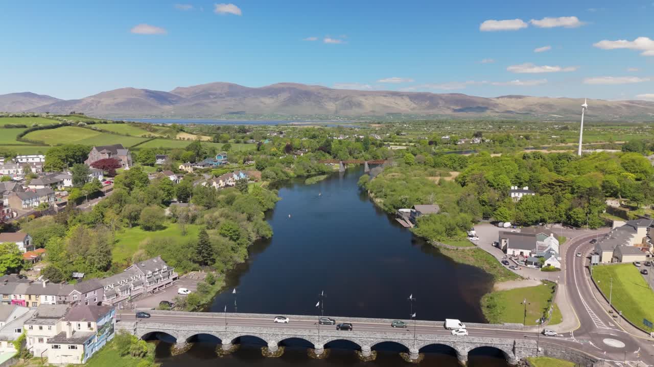 Drone over Killorglin on a sunny day, showing the river, bridge, charming townhouses, lush trees, windmill, and rolling Kerry hills in the distance.