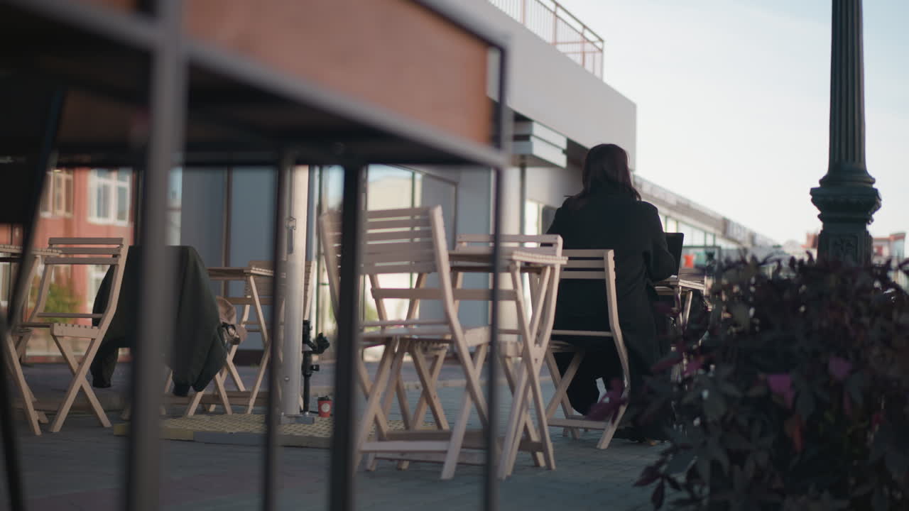 vista trasera de la dama con abrigo negro sentada en la mesa de la cafetería, escribiendo en la computadora portátil, su cabello se balancea suavemente con el viento en un entorno urbano pacífico, plantas y sillas de madera alrededor