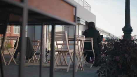 back view of lady in black coat seated at cafe table, typing on laptop, her hair sways gently with the wind in a peaceful urban setting, plants, and wooden chairs around