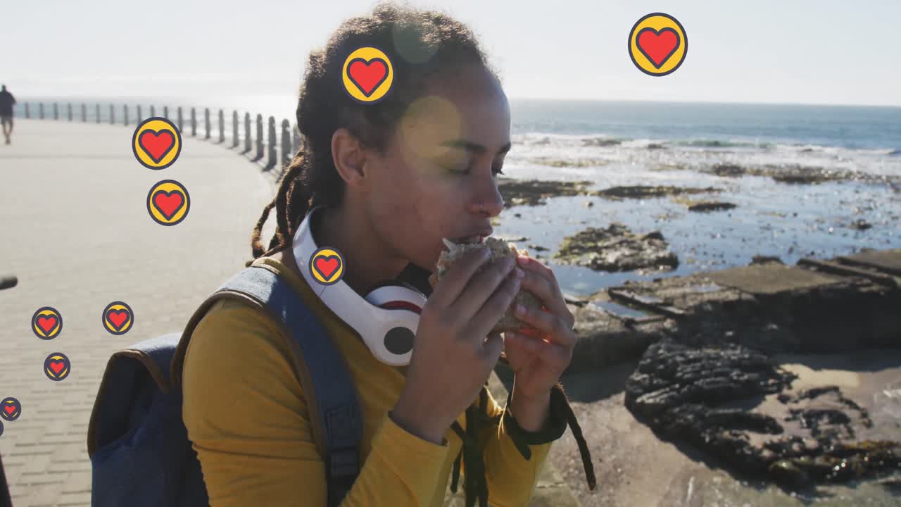 Multiple heart icons floating against african american woman eating a snack on the promenade