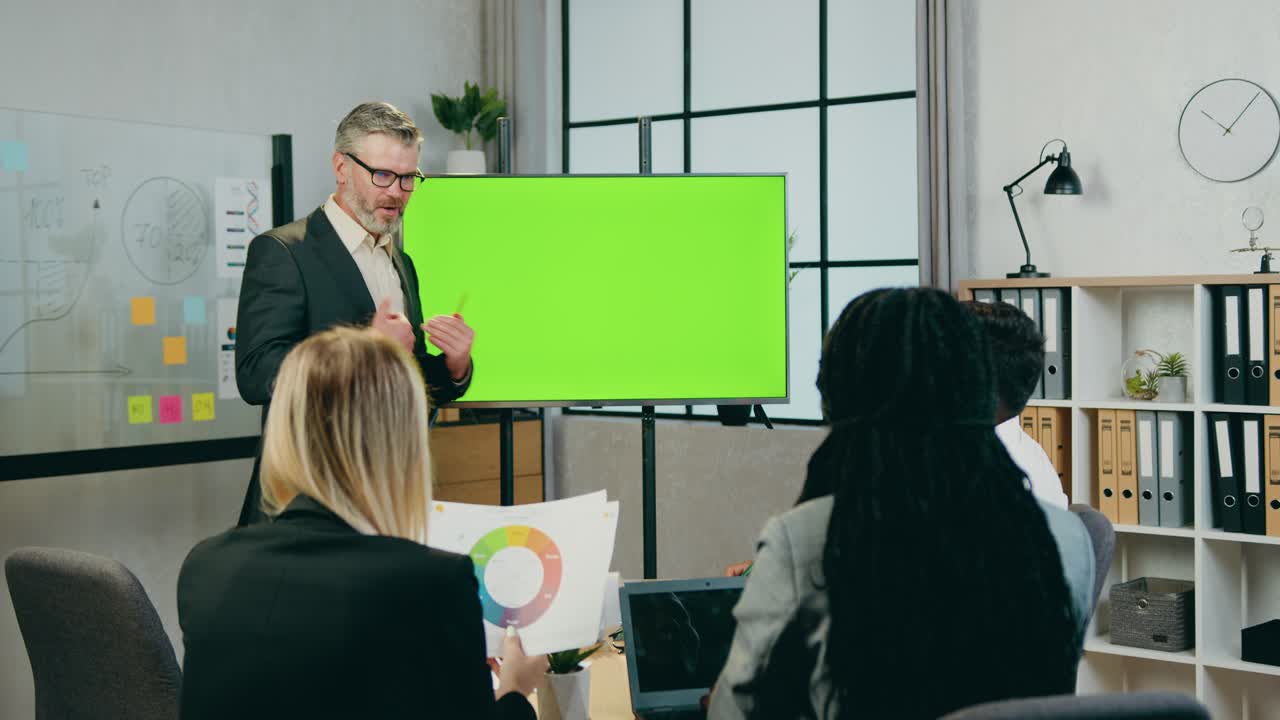 Attractive confident experienced bearded businessman talking with successful mixed race male and female colleagues while he standing near green screen in boardroom
