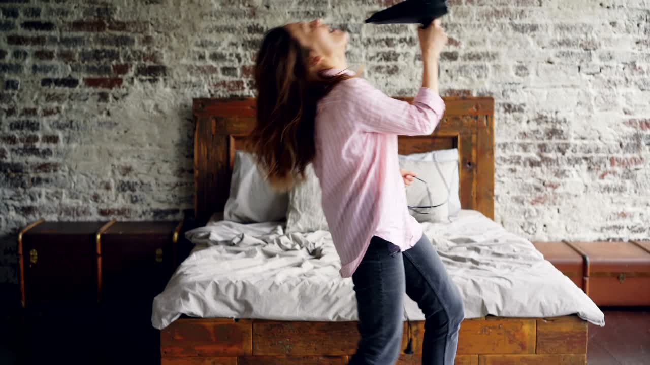 una joven atractiva con camisa y vaqueros está bailando y cantando sosteniendo un secador de pelo y usándolo como micrófono en un bonito dormitorio de estilo loft cerca de una cama doble moderna.