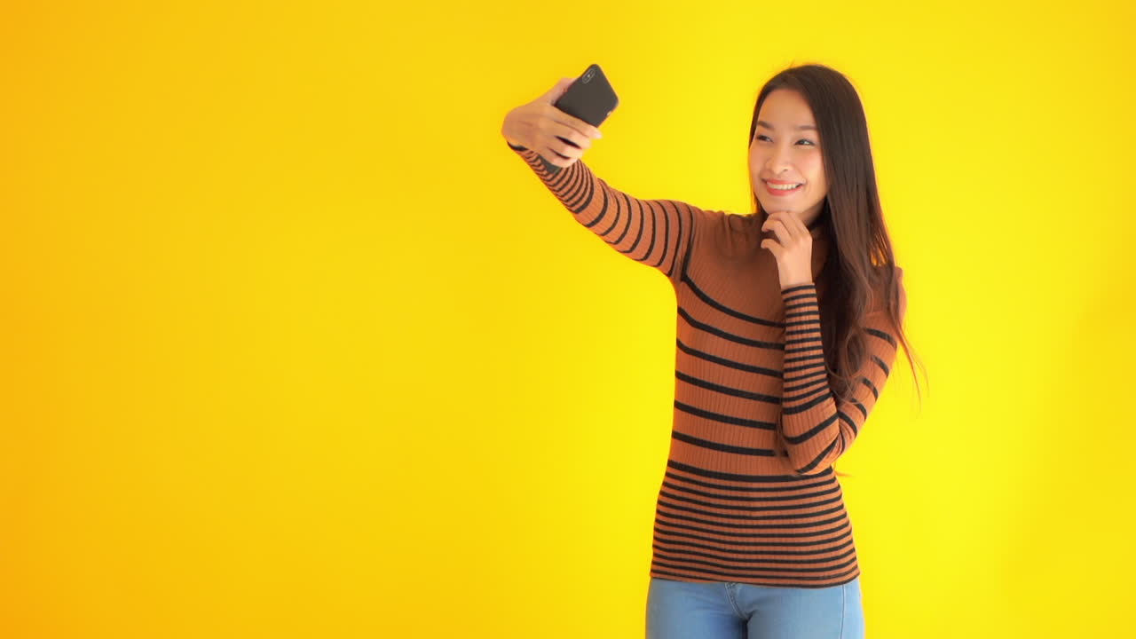 Static shot of a cute Asian lady smiling and taking a selfie in front of a yellow background