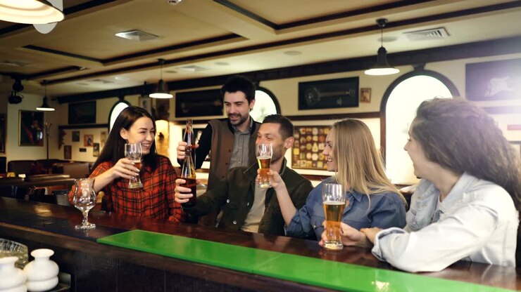 Students are drinking in bar celebrating end of academic session. Happy and sociable young people are chatting, toasting, clanging glasses and bottles and drinking.