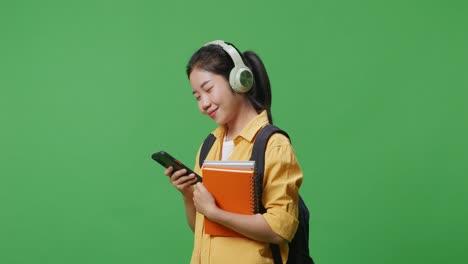 side view of smiling asian woman student with a backpack and some books wearing headphones and using a smartphone while standing in the green screen background studio