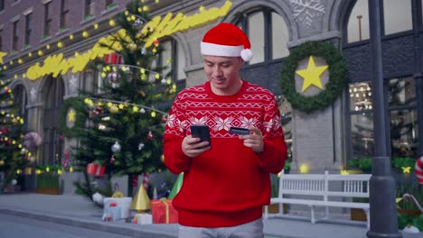 asian man in christmas suit with a smile holding a credit card and using smartphone spend money while standing in christmas night