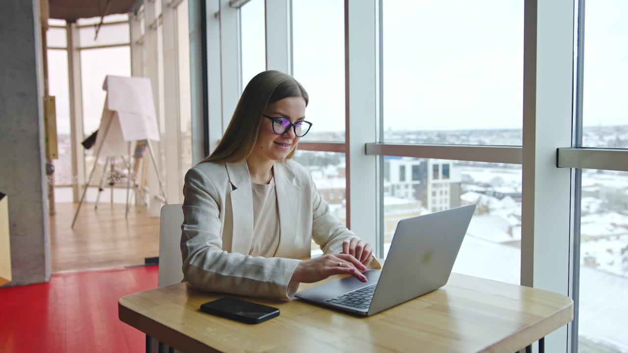 Smiling business lady sitting at the desk and working at laptop. Attractive woman at light office workplace. Panoramic windows with cityscape at backdrop.