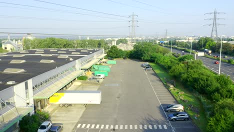 drone-moving-forward-along-a-small-warehouse,-along-the-highway-and-under-power-lines,-beautiful-weather