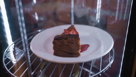 cheesecake slice topped with fresh strawberry slices placed on white plate inside glass display with reflections and wire shelf. creamy texture and golden top contrast beautifully with red fruit garnish