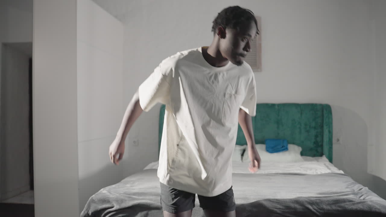 Man in white t-shirt holding backpack, standing next to bed with green headboard in minimalist bedroom, preparing to leave, sunlight coming through window, simple, clean decor in background