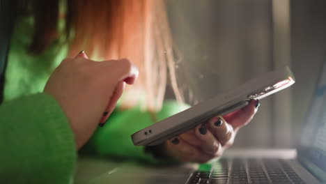 a woman in a green sweater holding and clicking on her smartphone while a laptop is in front of her. captured with a handheld camera, emphasizing multitasking and modern digital workspace