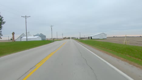 pov conduciendo por una carretera rural pasando por campos vacíos y tranquilos corrales en un día nublado de invierno en la zona rural oriental de lowa