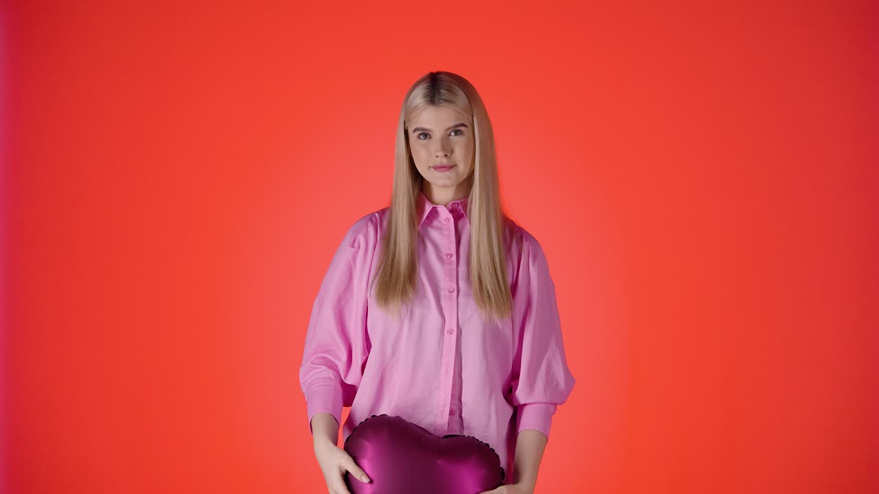 Bella donna bionda che lancia palloncini a forma di cuore viola in aria su sfondo rosso, scatto in studio.