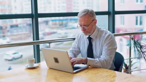 senior freelancer using laptop working from home. smiling grey-haired man in glasses typing on computer and drinks coffee enjoying distant job.