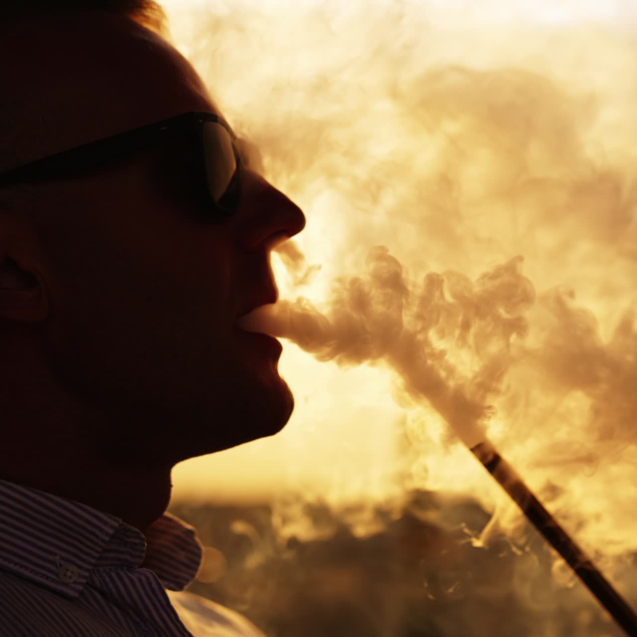 Silhouette of man in glasses smoking hookah sitting outdoors. Close up. Backdrop of setting sun