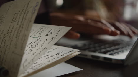 an asian woman types on her laptop keyboard out of focus while a notebook with notes flaps in the wind