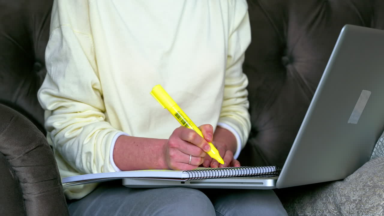 Caucasian woman sitting on a chair, working on her laptop and making notes in notepad