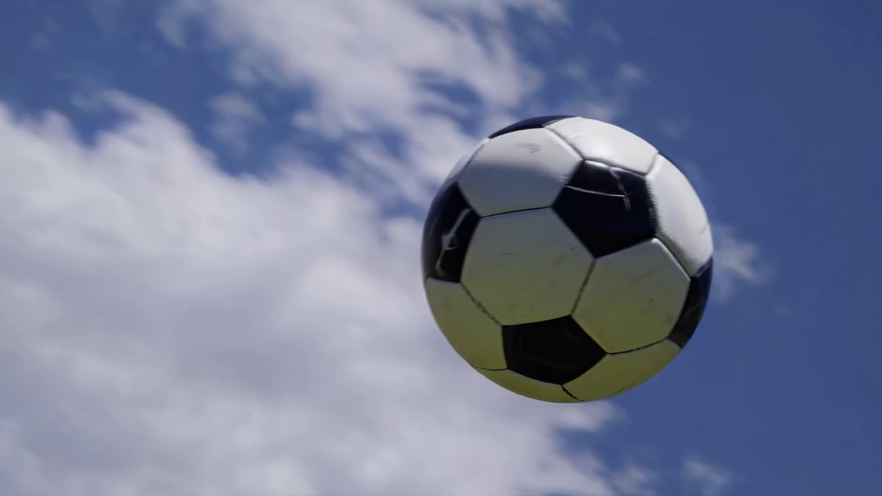 Inquadratura dal basso di un pallone da calcio a mezz'aria contro un cielo blu con nuvole, che cattura il movimento dinamico.