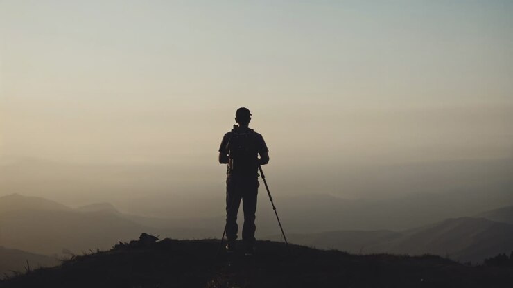 Escursionista che si sistema sulla cresta, inquadrando la luce dell'alba che arriva su una valle nebbiosa, posizionando una fotocamera su treppiede, berretto e zaino.
