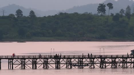 Mon-Bridge-and-a-longboat-followed-speeding-behind-the-structure-while-people-are-walking-on-the-bridge,-silhouetting-as-it-was-getting-dark,-in-slow-motion