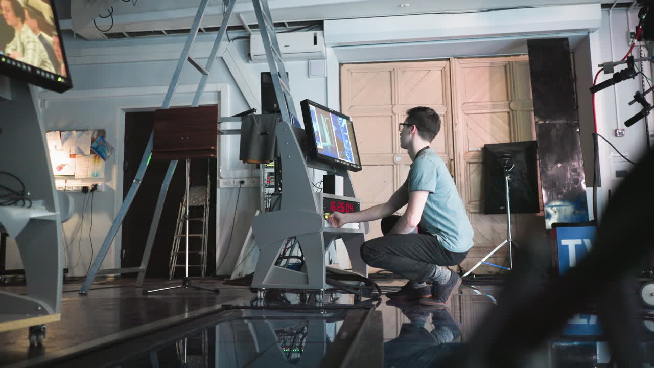 Man adjusting settings on digital equipment in modern studio, crouching near high-tech monitor and timer, professional production in process, technology-focused environment, action shot