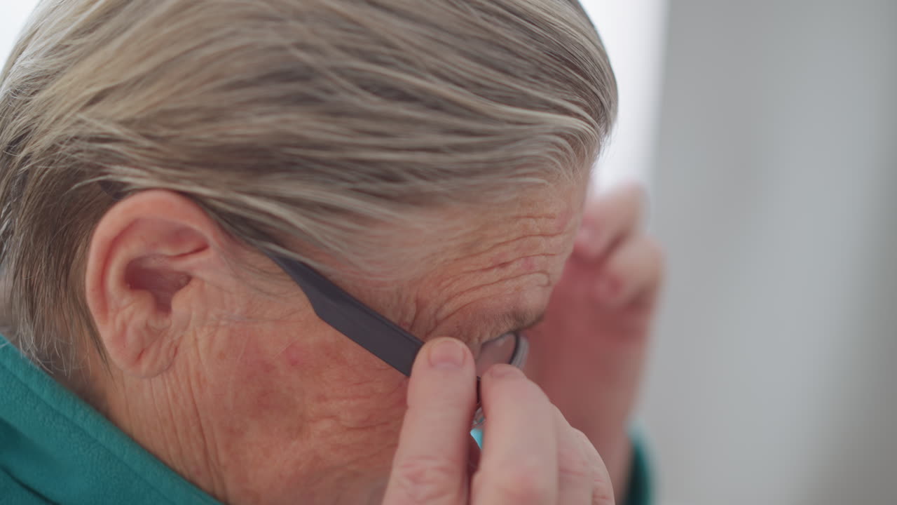 Lonely pensioner in green teal sweater sitting and adjusting glasses with both hands, closely examining glasses, engaged in daily activity at home, emphasizing her hands and focus
