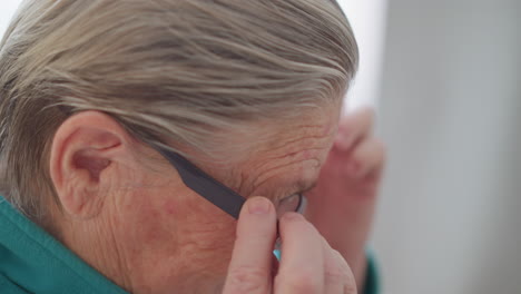 lonely pensioner in green teal sweater sitting and adjusting glasses with both hands, closely examining glasses, engaged in daily activity at home, emphasizing her hands and focus