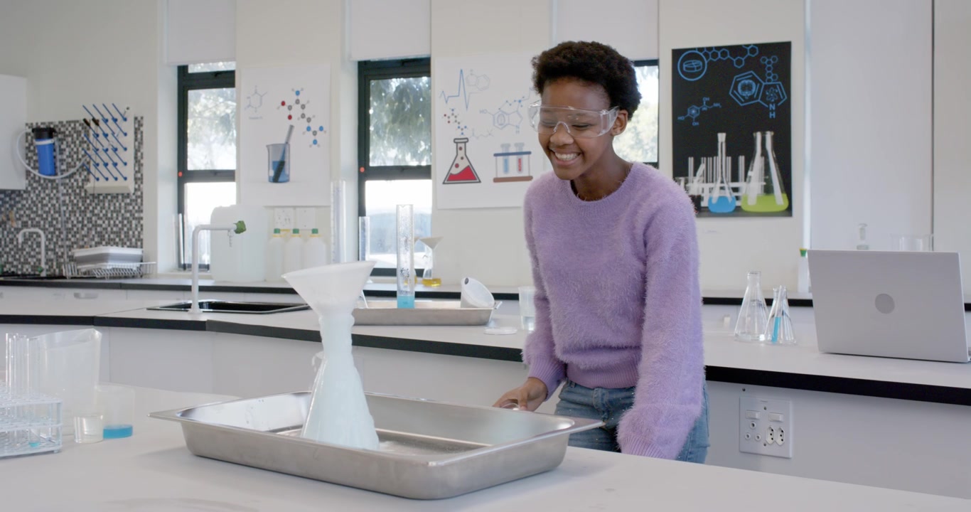 Teen African American doing chem demo in school lab pouring into funnel with goggles causing foam